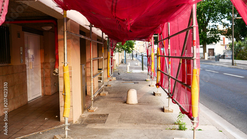 Red safety debris netting on scaffolding over city sidewalk. Construction site enclosure for pedestrian protection during building facade renovation, restoration and urban maintenance works outdoor.