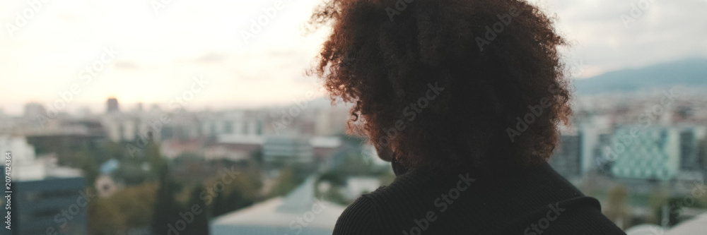 Fototapeta premium Close-up of young guy is standing on the balcony looking at the city, back view. Panoramic