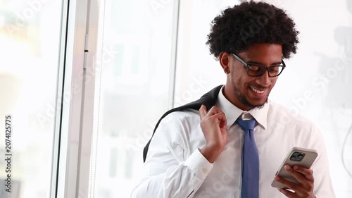 Young African American man looking at and typing on his cell phone, smiling, in a bright office by the window. Perfect for business, tech, and communication concepts