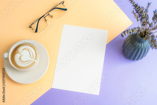 Blank paper mockup with coffee cup, eyeglasses and lavender flowers on purple and beige desk background. Top view, flat lay