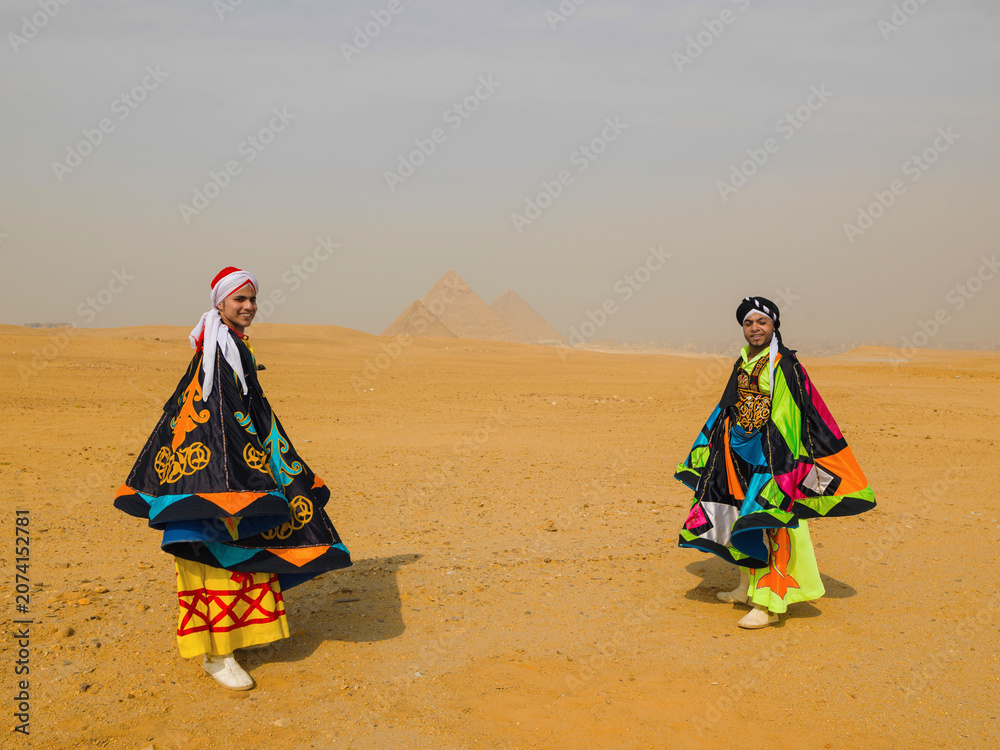 Fototapeta premium Two smiling Egyptian tanoura folk dancers in colorful traditional costumes performing in the desert before the Pyramids of Giza ideal for Egypt