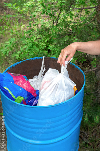 Hand placing a white plastic bag into a blue trash barrel outside.