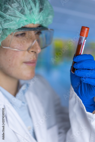 Person wears a white lab coat. Blue gloves cover their hands. They hold a test tube with red liquid. Safety goggles protect their eyes. Green hairnet secures their hair