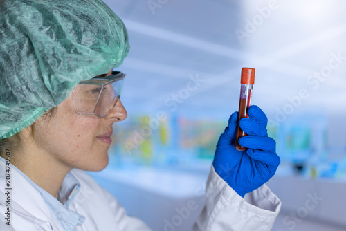 Lab technician wears green cap and blue gloves. They hold a test tube close to inspect. Close view shows precise laboratory work. Background has blurred lab shelves