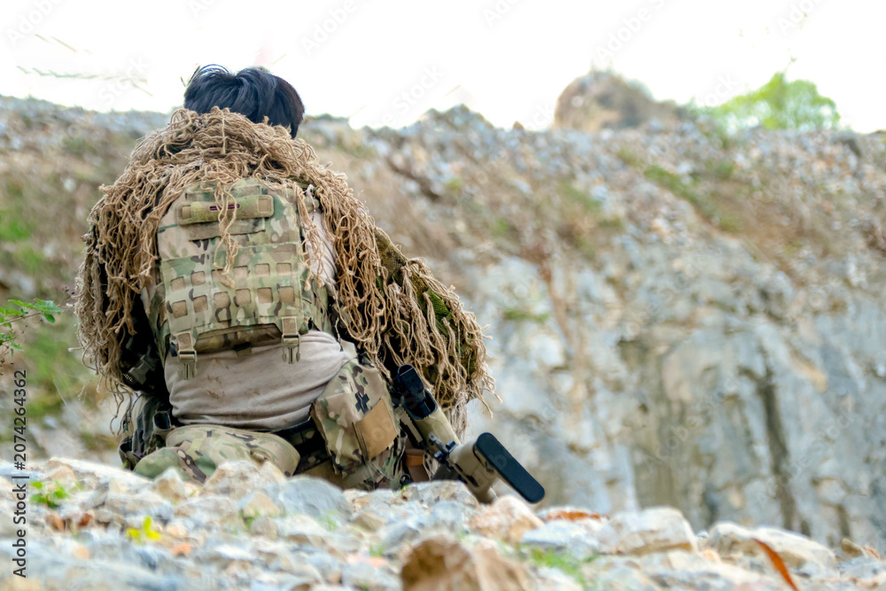 Fototapeta premium Rear view male soldier in camouflage uniform sitting on a rock on the edge high cliff in a mountainous area closely observing the border situation.