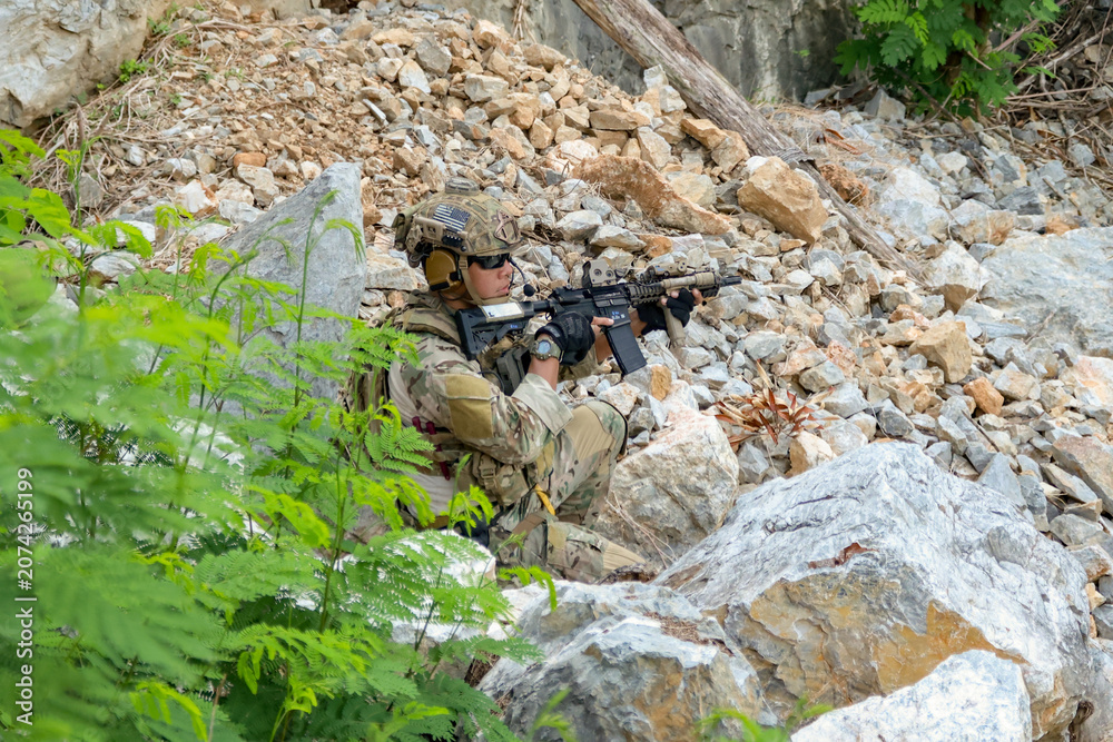Fototapeta premium Male soldier in camouflage uniform sits on a rock on the edge high cliff in a mountainous area closely monitoring the border security situation.