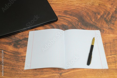 open squared notebook with a black pen on a brown wooden table near the edge of a laptop