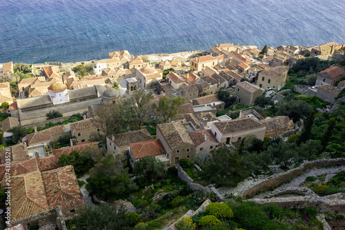 Photography A high-angle view of the historic town of Monemvasia, Greece, with its stone houses and red-tiled roofs nestled on a rocky outcrop overlooking the deep blue Aegean Sea