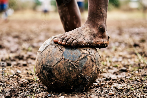 African child's muddy feet resting on a worn soccer ball on a dirt pitch