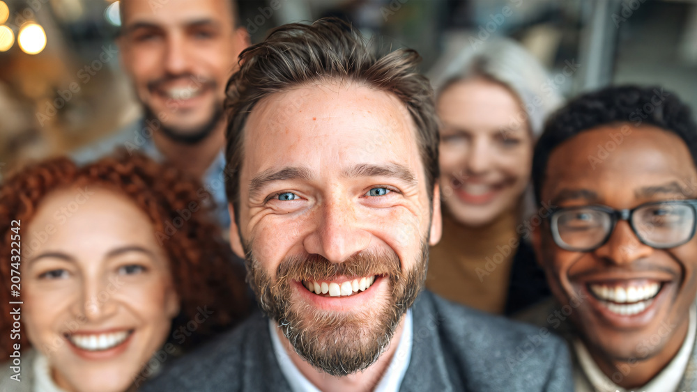 Obraz premium A smiling diverse business team gathered closely together in a modern office, looking upward toward the camera to represent unity, leadership, and successful collaboration.