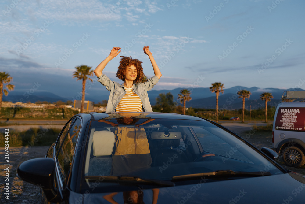 Fototapeta premium Young woman enjoys a joyful moment through an open car sunroof in a desert setting, casual denim jacket, striped top, warm sunset light, free spirited lifestyle
