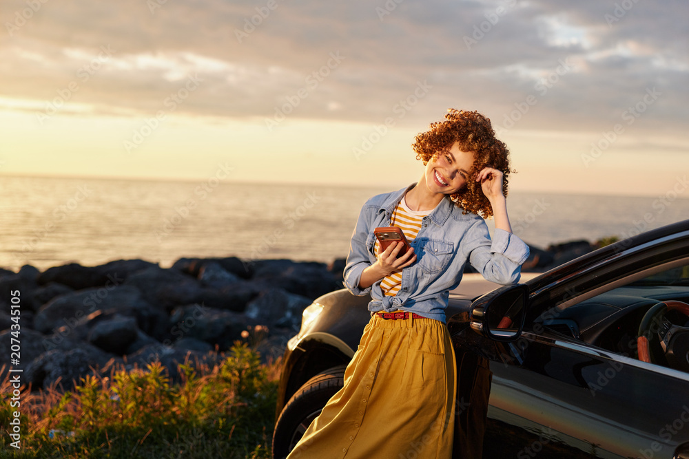 Fototapeta premium Young woman stands beside a car at sunset along the coast, talking on her phone with a relaxed candid mood, wearing a denim shirt and bright yellow skirt.