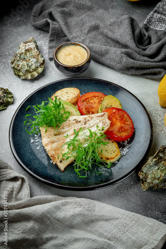Vertical restaurant food composition with grilled white fish, roasted vegetables, fresh herbs, lemon and oyster shells on grey textured background