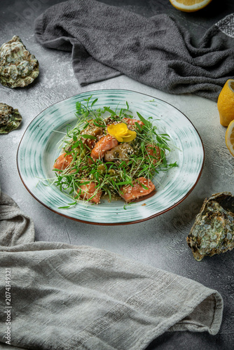 Vertical seafood salad with salmon, sesame seeds, microgreens, edible flower, lemon and oyster shells on textured grey restaurant table
