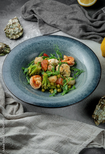 Vertical seafood restaurant dish with shrimp, scallops, broccoli, cauliflower, peas, tomato and fresh herbs on a grey textured table