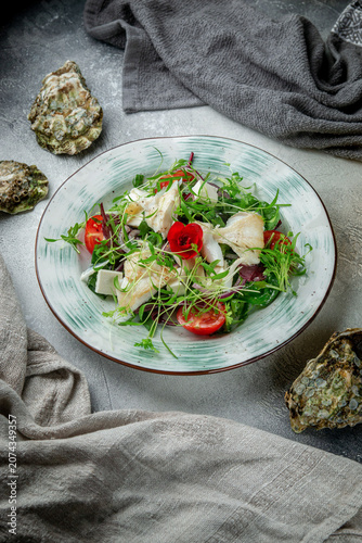 Vertical seafood salad with white fish, mixed greens, tomatoes, cheese, microgreens and oyster shells on textured grey restaurant table