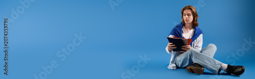 Fotografi Handsome young student with long hair immersed in reading against a vibrant blue