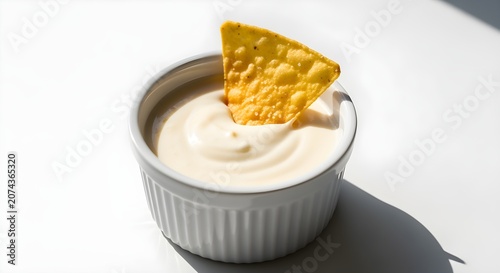 A small ramekin of smooth white queso with one tortilla chip dipping in, on white.