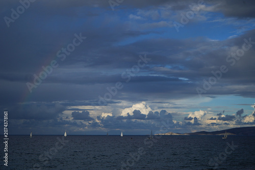 A fleet of sailboats on the horizon under a dramatic, stormy sky in Crete, with a faint rainbow appearing over the dark Mediterranean sea.
