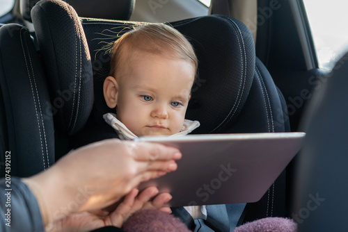 Valokuva Mother is giving a tablet to her toddler daughter sitting in car seat to enterta