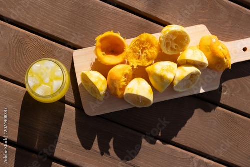 Overhead view of halved and squeezed lemons arranged on wooden cutting board beside glass of iced lemonade on slatted wood table, bright sunlight casting