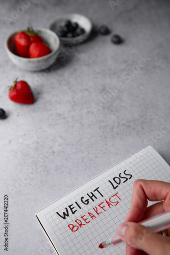 A hand writing Weight loss breakfast in a notebook next to healthy berries and ingredients.