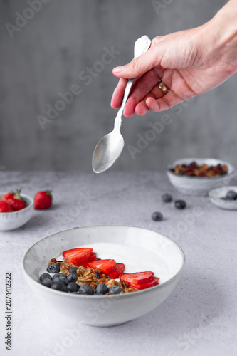 A woman's hand holding a spoon over a yogurt bowl with granola and fresh berries