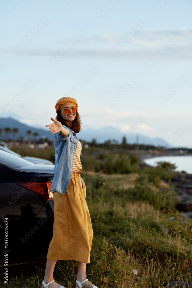 Fototapeta premium Woman lifestyle outdoors by coast, standing near car, sunny day, relaxed pose, pointing to camera, hat, denim jacket, mustard skirt, striped top, natural scenery, tranquil mood, travel vibes