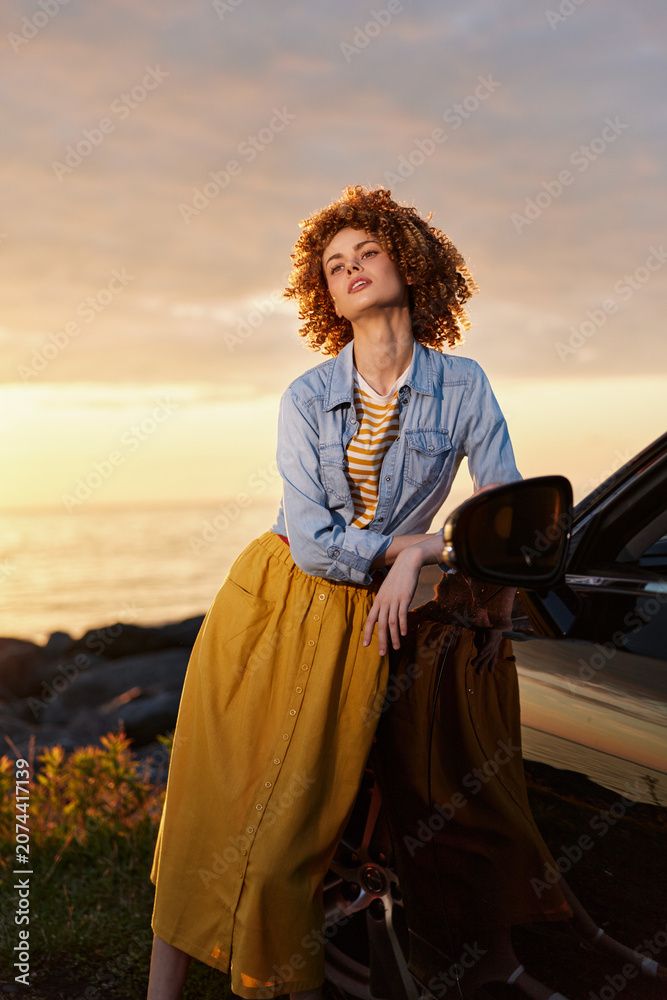 Fototapeta premium Relaxed woman with curly hair leaning against a car at sunset, wearing a denim shirt and mustard skirt. A candid lifestyle moment focused on fashion and outdoor confidence.