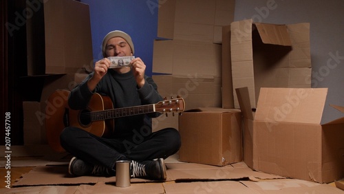 Fotografija Man in a beanie and sweater sitting with a guitar and a donation can, happily ho
