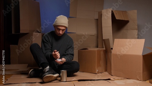 Bild auf Leinwand Young man wearing a beanie and dark clothing, sitting alone on cardboard, counti
