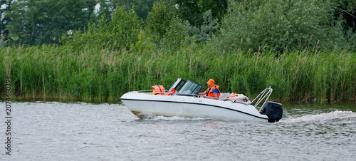 Small speedboat moves quickly on a lake past lush green shore vegetation. Perfect for themes of active lifestyle, weekend leisure, nature travel, and river navigation. Photo