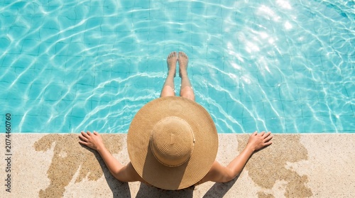 Top-down view of woman in wide straw sun hat sitting on pool edge, hands on concrete, feet in clear blue water with ripples and reflections