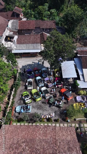 Aerial view of a gathering of vintage and classic cars parked outdoors in a village setting on a sunny day