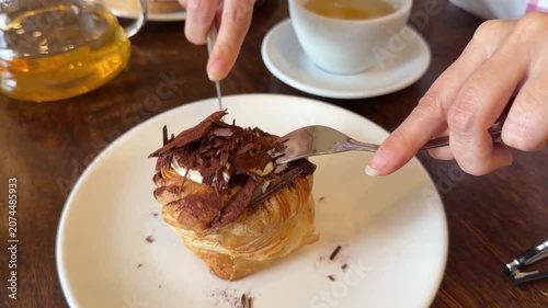 Fresh laminated pastry topped with chocolate cream and dark chocolate shavings is cut with fork, knife before serving at cafe table with tea. Sweet dessert, bakery, food indulgence, dining experience