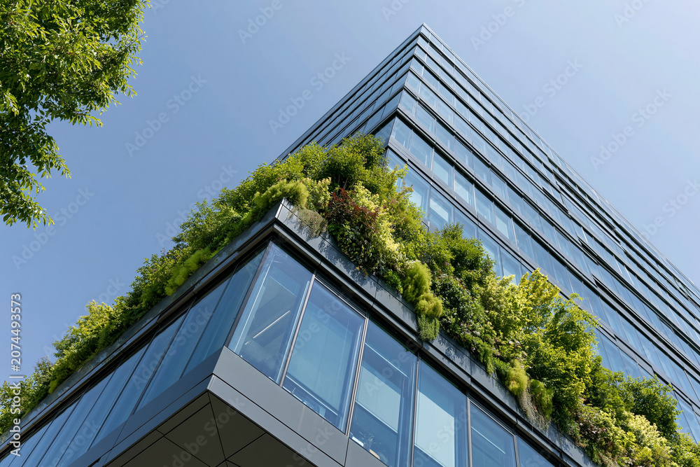 Fototapeta premium Modern building features lush green terraces. Glass and steel reflect bright blue skies. Vibrant foliage climbs vertical surfaces. Urban architecture blends nature harmoniously