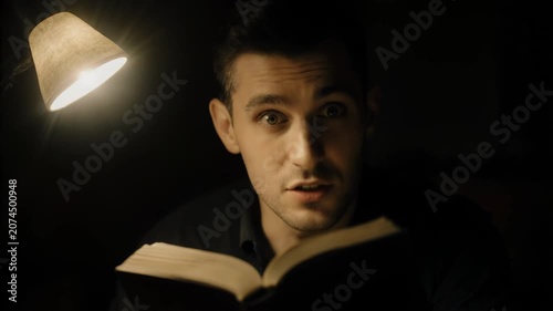 Male reader with short dark hair engaged in a book under warm lamp light, showing expressions of surprise and curiosity in a dimly lit indoor setting