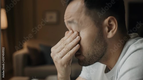 Male individual with glasses sits at a table, showing visible signs of stress while holding his head in a cozy living room with soft lighting and comfortable furniture