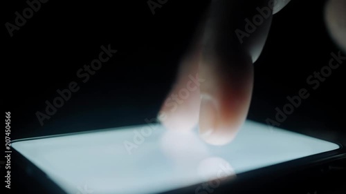 Close-up of a finger tapping a smartphone screen, showcasing the interaction with the illuminated display in a dark environment, highlighting the touch action and device details