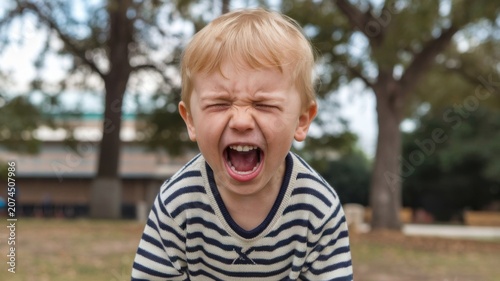 Young boy screams with eyes shut tight and mouth wide open. Child has blond hair, wears striped jumper. Toddler displays strong emotion outdoors.