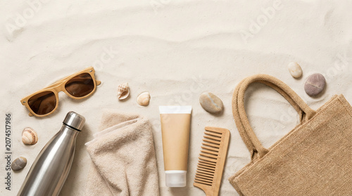 Summer beach essentials arranged on sand, featuring sunscreen, sunglasses, towel, reusable bottle, and tote bag in a minimal travel flat lay composition. copy space