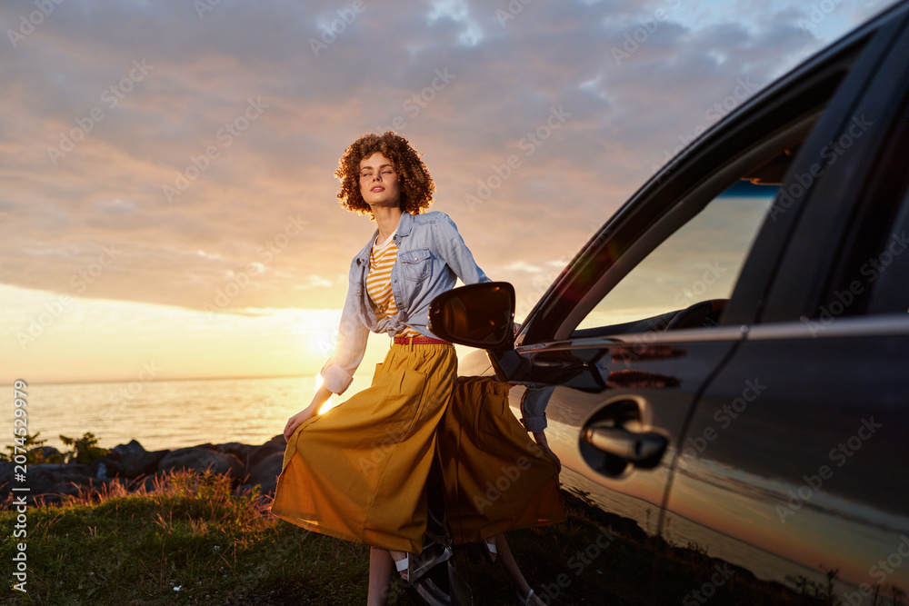 Fototapeta premium Woman by a car at sunset, wearing a denim shirt and yellow skirt, enjoying a carefree outdoor lifestyle moment, breeze in hair, warm golden light, posed near a scenic coast, soft shadows, candid