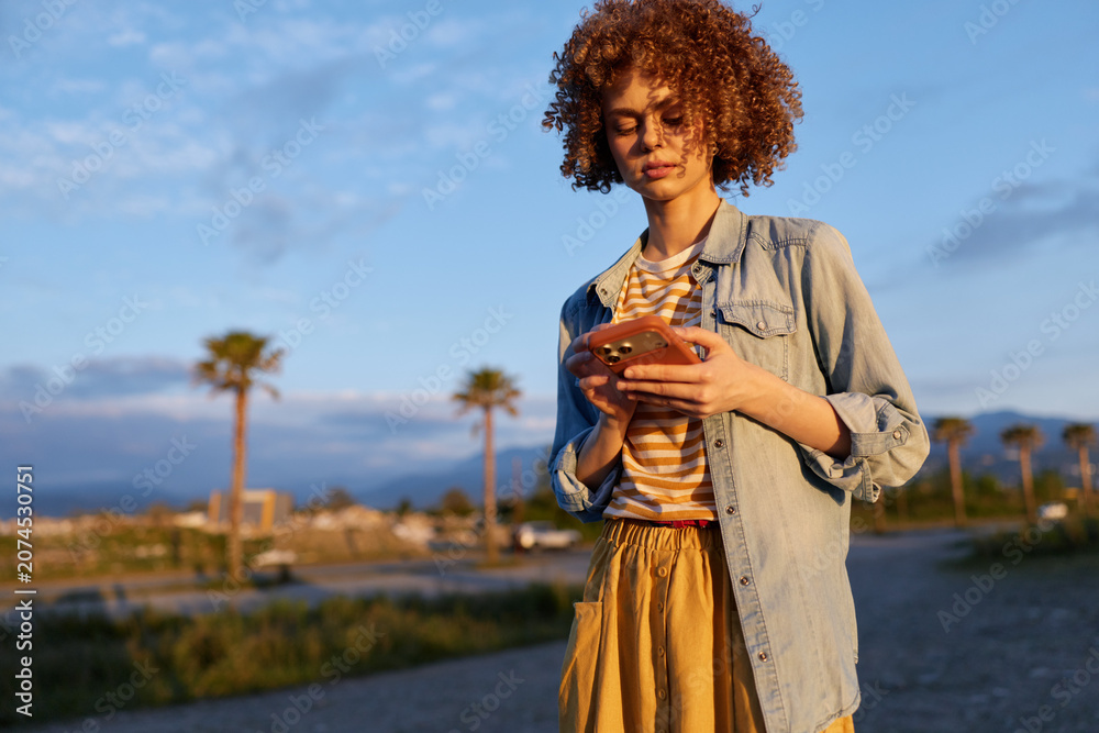 Fototapeta premium Woman on a sunny outdoor street uses smartphone, casual denim shirt over striped tee, flowing skirt, palm trees in background, candid lifestyle moment, modern relaxation