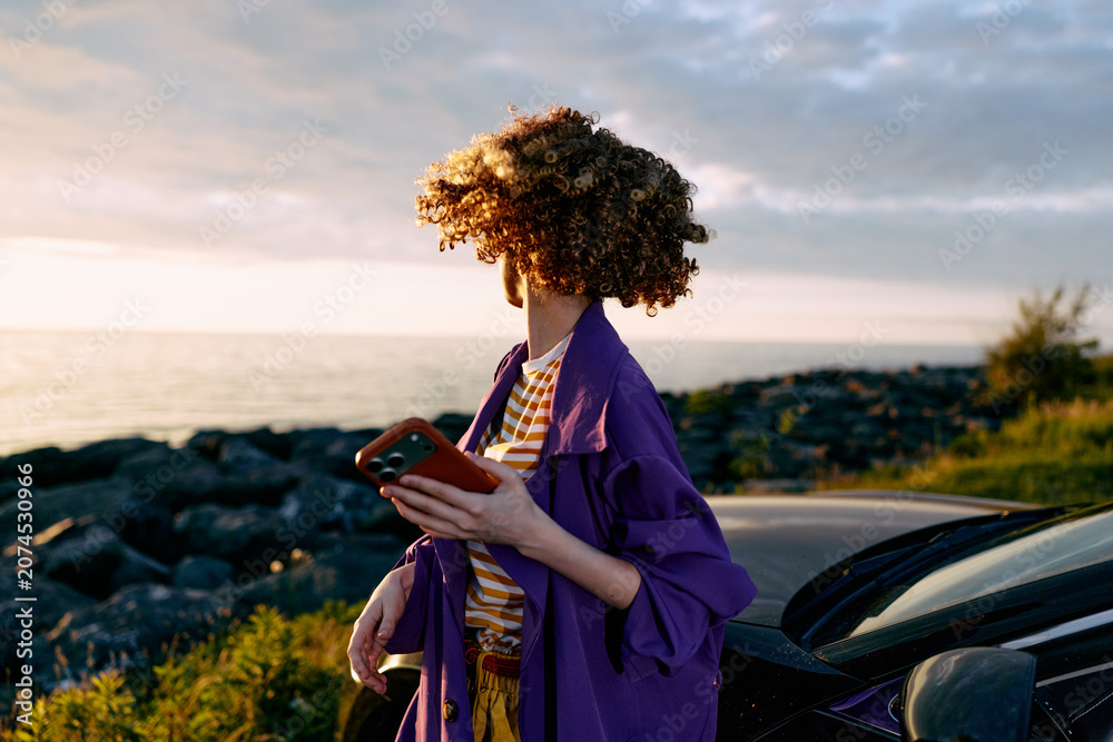 Fototapeta premium A woman stands by a car on the coast at sunset, her hair blowing in the breeze, looking away while holding a tablet. A lifestyle moment of independence and travel.