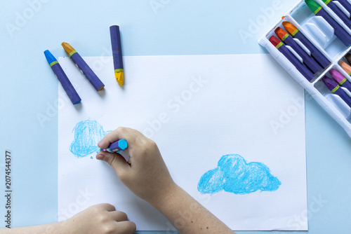 Top-down view of a child's hands drawing blue clouds with oil pastels on white paper, scattered purple crayons and a white supply box on a light blue table, creative art activity.