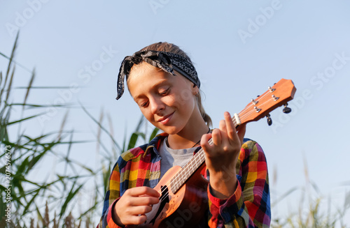 Happy teen girl playing ukulele guitar outdoor at sunset. Smiling teenage female musician in plaid shirt and bandana enjoying music hobby in summer field