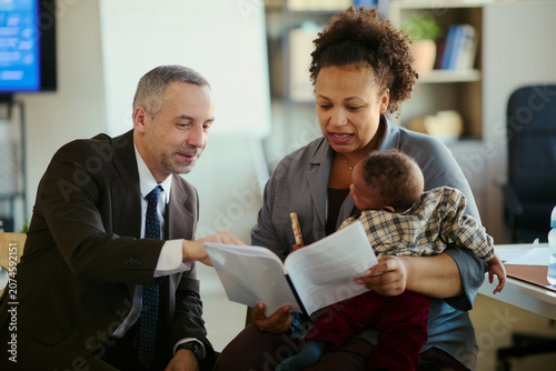 Фотография African American mother holding baby while reviewing documents with middle aged Caucasian businessman in office, showing working parent support