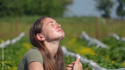 Profile portrait of a young female farmer tasting a fresh ripe strawberry right on a sunny plantation field.