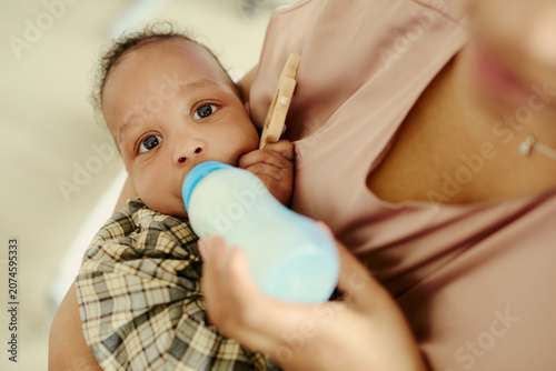 Fotografie Portrait of Black infant drinking from baby bottle while mother holding child close in workplace, showing working motherhood and childcare balance