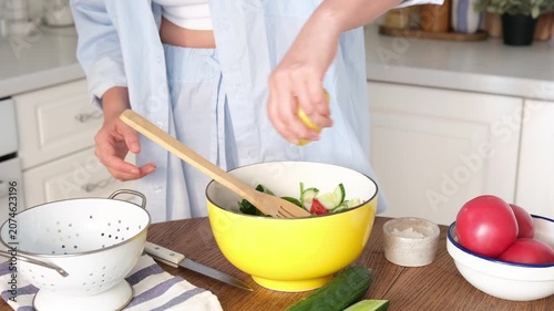 Cropped person squeezes lemon over salad in yellow bowl inside bright home kitchen, with hands centered above mixed vegetables, wooden spoon resting in bowl, and clean daylight around tabletop